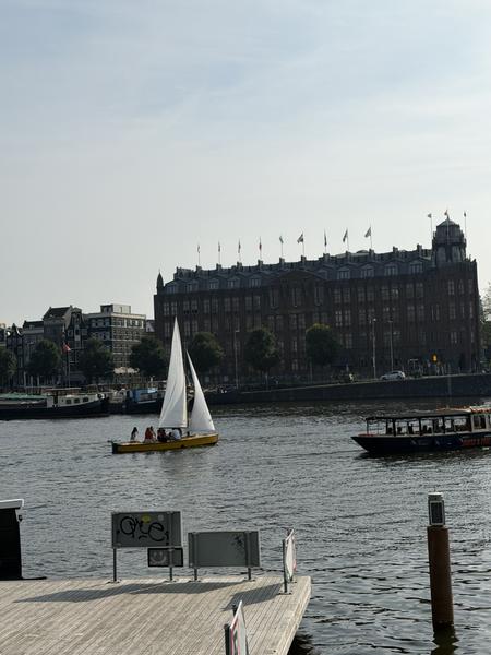 This photo captures a lively scene on the water at Oosterdoksstraat, Oosterdokseiland, Amsterdam, with a yellow sailboat and a larger canal boat cruising past a grand historic building